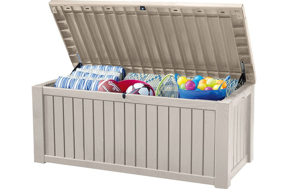 Kremowa skrzynia ogrodowa z otwartą pokrywą, wypełniona różnymi przedmiotami, w tym piłkami i kocami. Skrzynia ma pionowe żłobienia na zewnętrznej powierzchni.