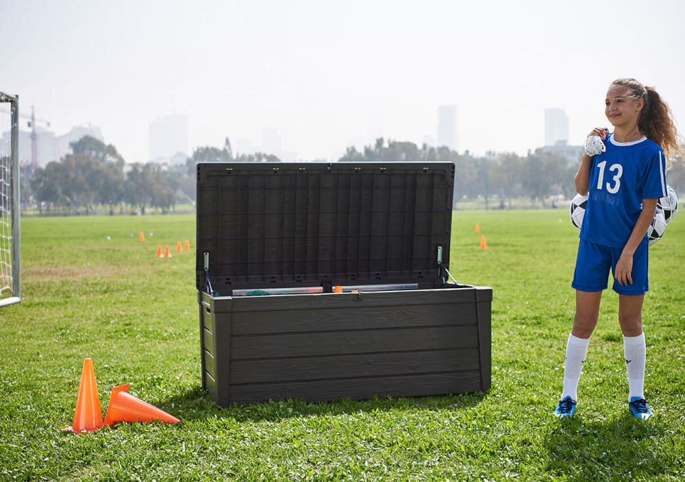 Na trawniku stoi otwarta skrzynia ogrodowa w ciemnym kolorze, obok której znajduje się osoba w niebieskim stroju sportowym z numerem 13. W tle widoczne są drzewa i zarysy budynków, a na trawie leżą pomarańczowe pachołki.