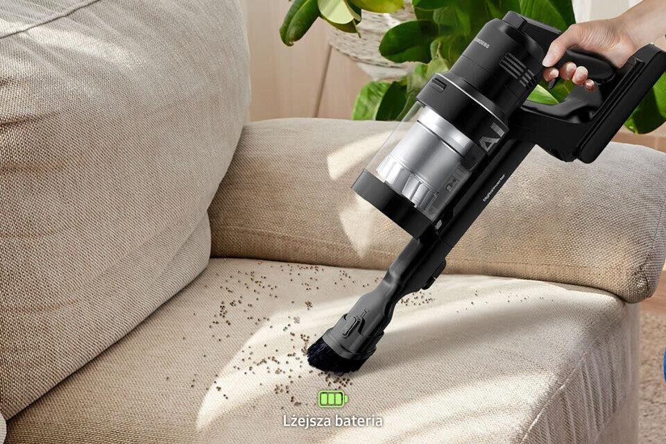 Osoba używa odkurzacza do czyszczenia kanapy z rozsypanych drobinek. Na dole obrazu widoczny jest tekst: \'Lżejsza bateria\'.