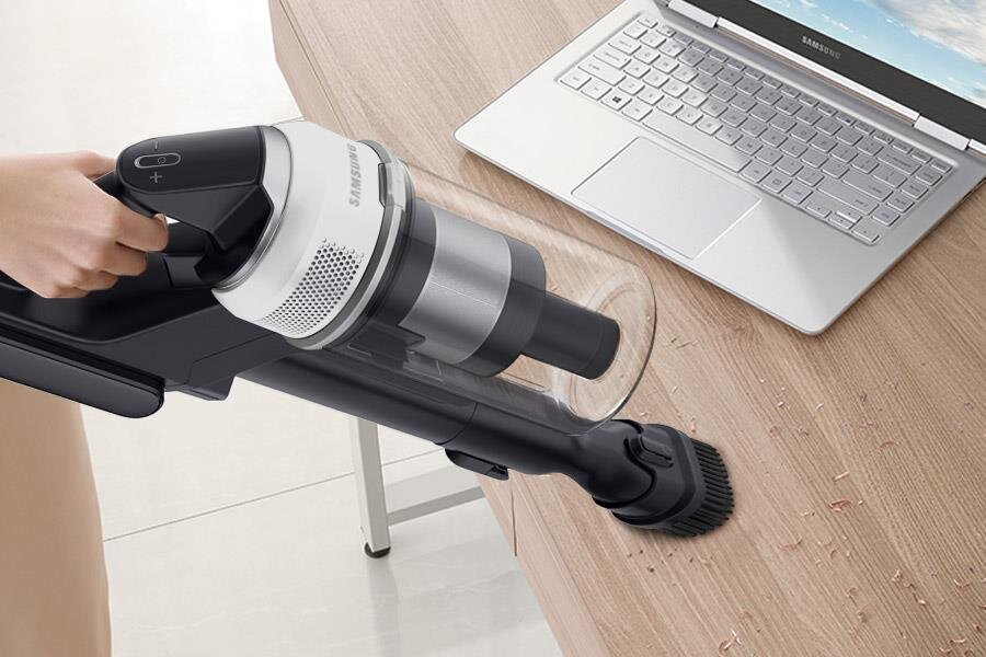 Osoba używa odkurzacza ręcznego do czyszczenia blatu stołu, na którym znajdują się okruchy. Obok znajduje się otwarty laptop z widocznym logo Samsung.