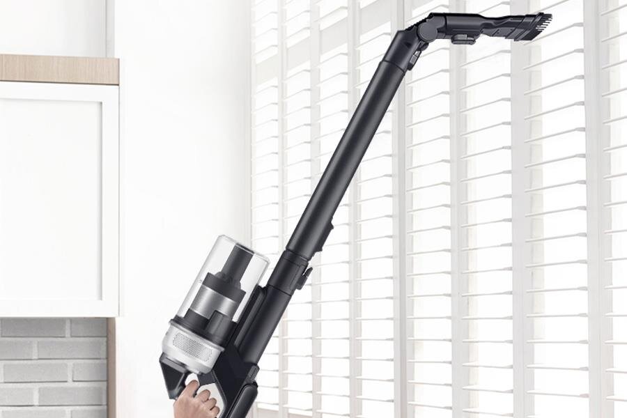 Osoba trzyma odkurzacz z długą, czarną rurą, skierowany w stronę białych żaluzji. W tle widoczna jest część kuchni z białą szafką i ceglaną ścianą.