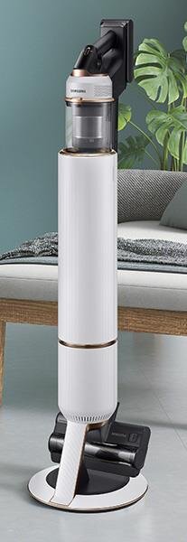 Na obrazie widoczny jest biały odkurzacz pionowy z przezroczystym pojemnikiem na kurz, stojący na okrągłej podstawie. W tle znajduje się fragment kanapy oraz roślina doniczkowa.