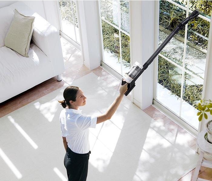 Osoba używa odkurzacza pionowego w jasnym pomieszczeniu z dużymi oknami. W tle widać białą sofę i rośliny za oknem.