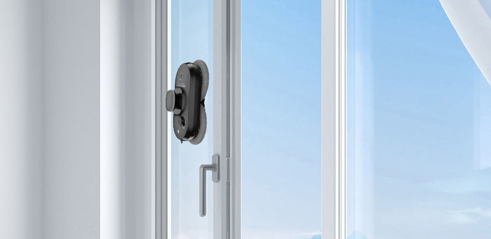 Na szklanej powierzchni okna znajduje się czarny robot do mycia okien. W tle widać jasne niebo i fragment zasłony.