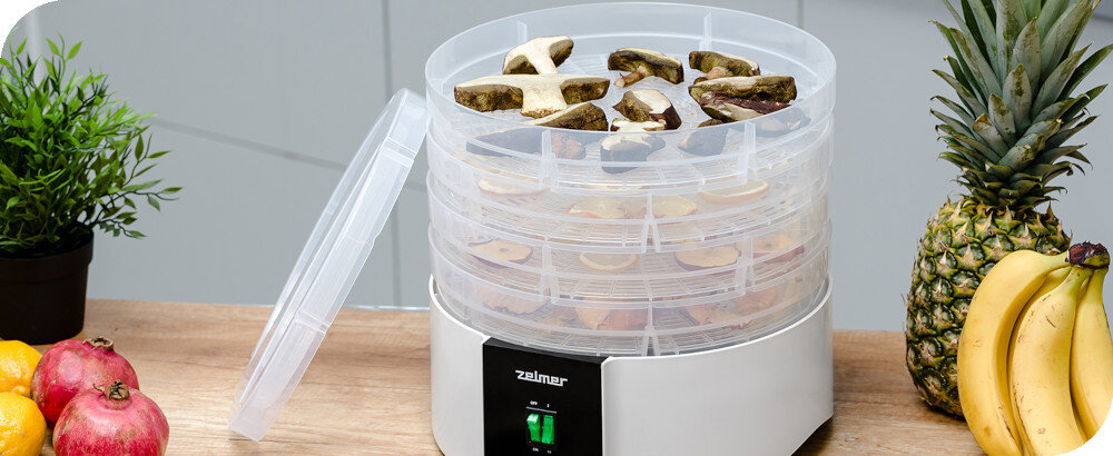 Na blacie kuchennym stoi suszarka do grzybów z przezroczystymi tackami, na których znajdują się kawałki grzybów. Obok suszarki leżą owoce, takie jak ananas, banany i granaty, a na urządzeniu widoczne jest logo \'zelmer\'.