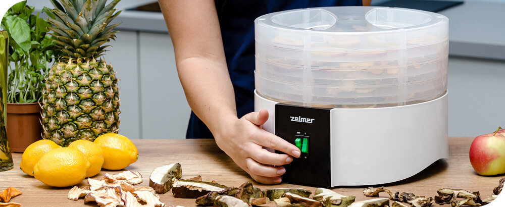 Na blacie kuchennym stoi suszarka do grzybów z logo \'zelmer\', obok której leżą owoce i kawałki grzybów. Osoba obsługuje urządzenie, naciskając przycisk na panelu.