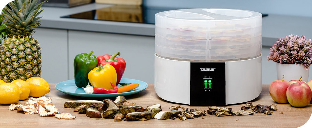 Na blacie kuchennym stoi suszarka do grzybów z logo \'zelmer\', obok której znajdują się owoce, warzywa i pokrojone grzyby. W tle widoczny jest fragment kuchni z płytą grzewczą.