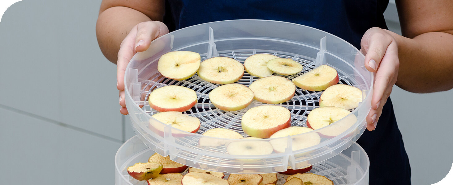 Osoba trzyma przezroczystą tacę z plasterkami jabłek, umieszczoną na suszarce do żywności.