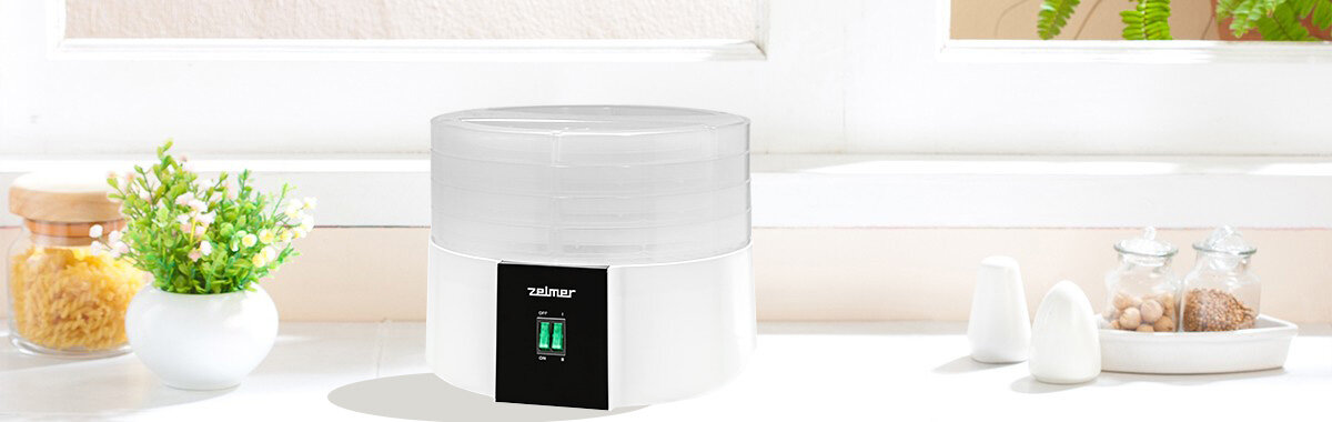 Na białym blacie stoi suszarka do grzybów z przezroczystą pokrywą i czarnym panelem z logo \'zelmer\'. W tle widoczne są rośliny doniczkowe i przyprawy w szklanych pojemnikach.