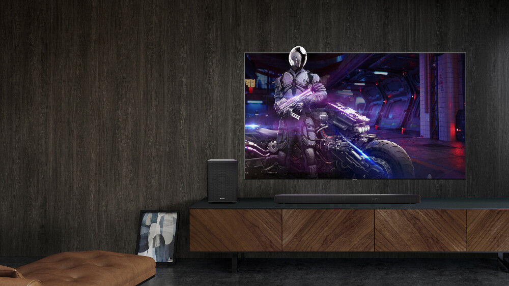 Na obrazie znajduje się nowoczesny salon z dużym telewizorem wyświetlającym postać w futurystycznym stroju. Przed telewizorem umieszczony jest soundbar oraz subwoofer na drewnianej szafce.