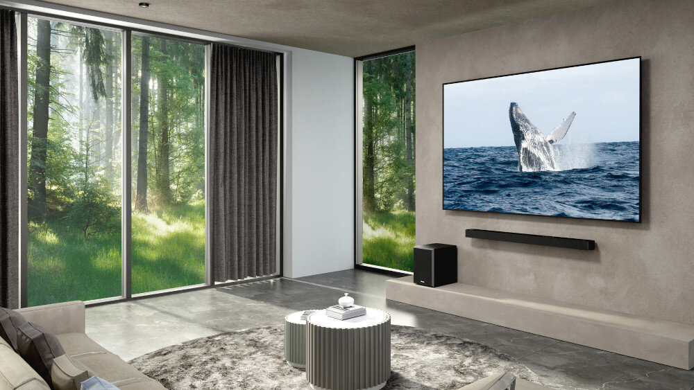 W nowoczesnym salonie znajduje się duży telewizor z obrazem skaczącego wieloryba nad wodą. Pod telewizorem zamontowany jest soundbar, a obok stoi bezprzewodowy subwoofer.