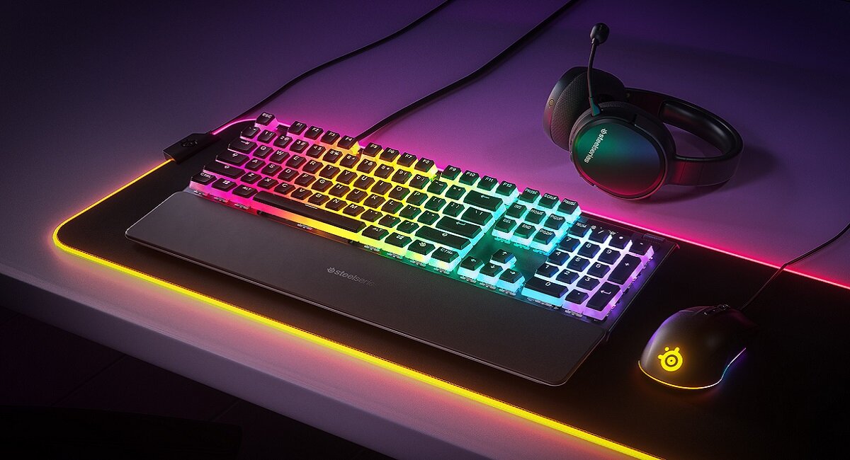 Na biurku znajduje się klawiatura z podświetleniem RGB, myszka oraz słuchawki. Na podkładce pod mysz widoczne jest logo SteelSeries.