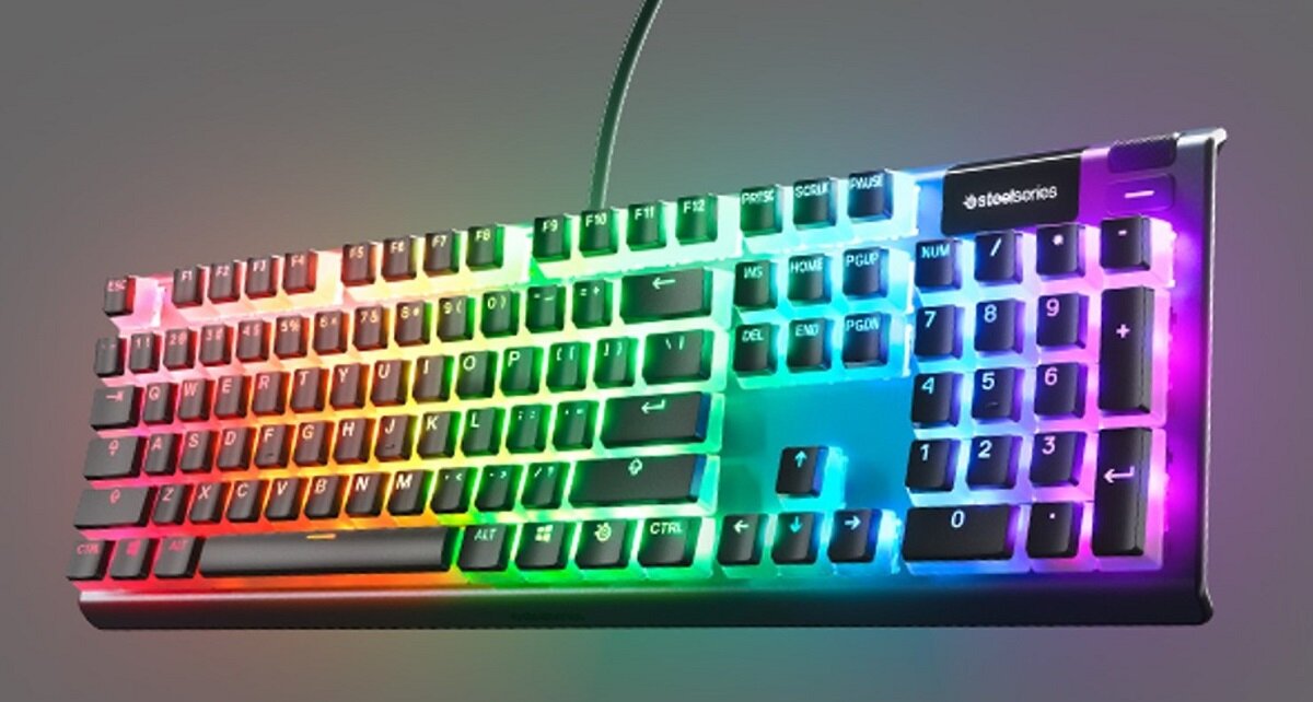 Klawiatura z podświetleniem RGB, z widocznym logo \'steelseries\' w prawym górnym rogu. Klawisze mają różnokolorowe podświetlenie, tworząc efekt tęczy.