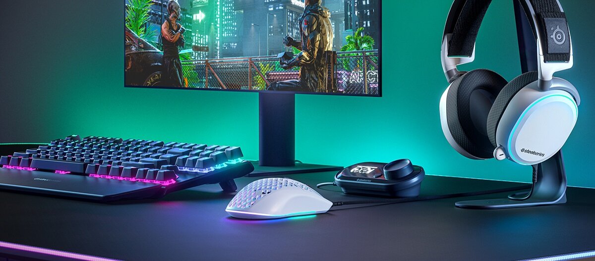 Na biurku znajduje się klawiatura z podświetleniem RGB, mysz, słuchawki na stojaku oraz monitor z grą. Na słuchawkach widoczne jest logo \'SteelSeries\'.