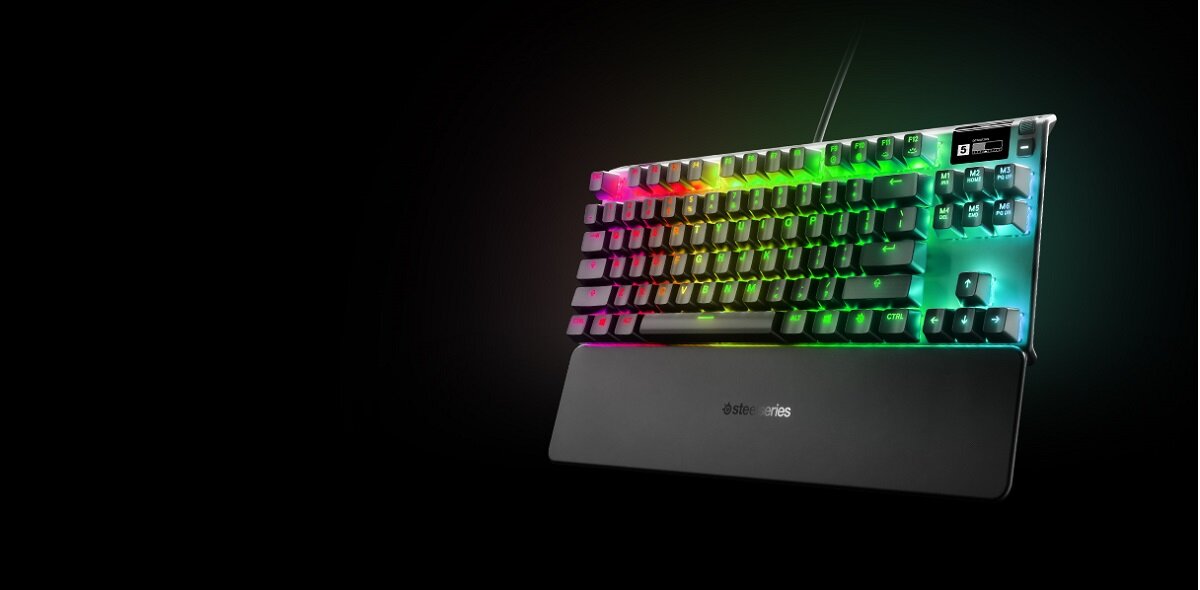 Na obrazie widoczna jest klawiatura z podświetleniem RGB, z logo \'steelseries\' na podpórce na nadgarstki. Klawisze są czarne, a podświetlenie mieni się różnymi kolorami.