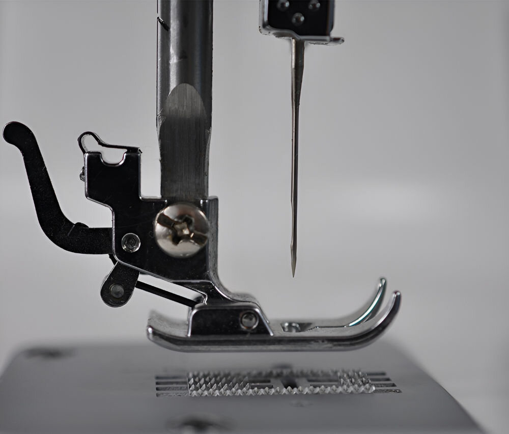Zbliżenie na stopkę maszyny do szycia z igłą skierowaną w dół. Widoczne są metalowe elementy konstrukcji na tle szarej powierzchni.