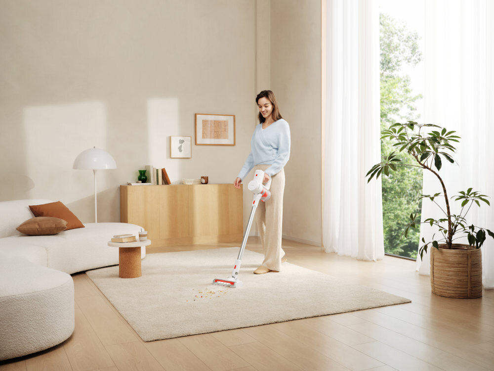 Osoba używa odkurzacza pionowego na jasnym dywanie w nowoczesnym salonie. W tle widać drewnianą komodę, roślinę w doniczce i duże okno z zasłonami.