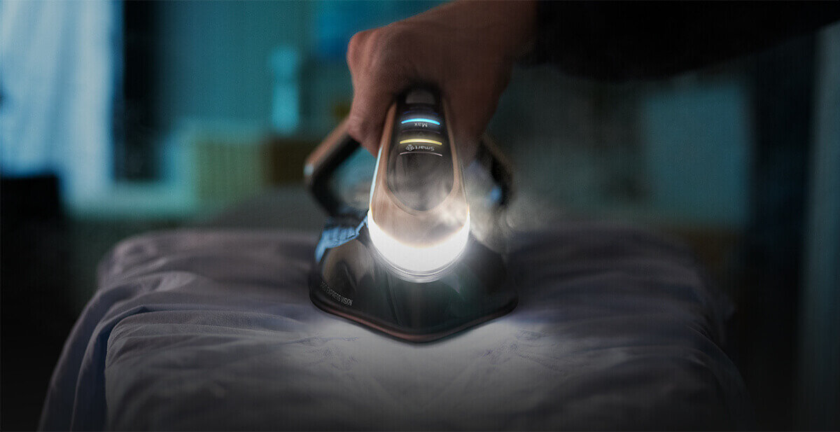 Osoba prasuje tkaninę za pomocą żelazka z oświetleniem LED, z którego wydobywa się para. W tle widoczne jest rozmyte wnętrze pomieszczenia.