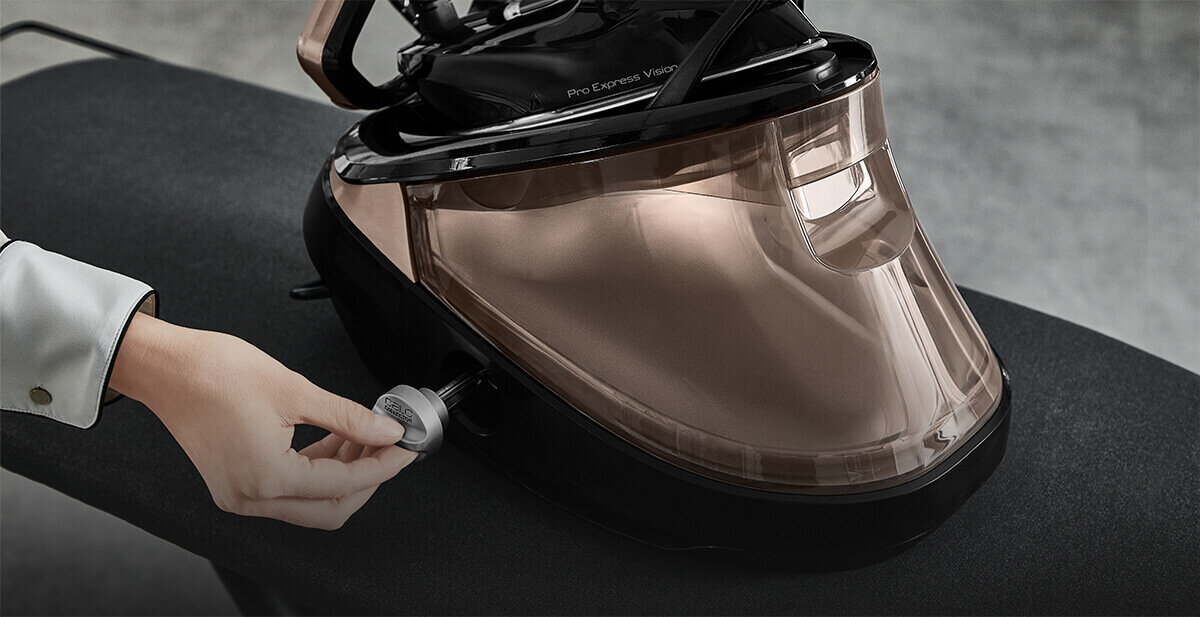 Osoba trzyma pokrętło przy generatorze pary z napisem \'Pro Express Vision\'. Urządzenie ma przezroczysty, brązowy zbiornik i znajduje się na ciemnej powierzchni.