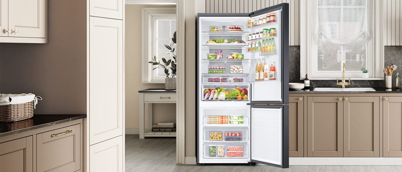 W kuchni znajduje się otwarta lodówka w kolorze grafitowej stali, wypełniona różnorodnymi produktami spożywczymi. W tle widoczna jest elegancka kuchnia z jasnymi szafkami i dużym oknem.