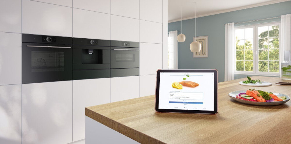 W nowoczesnej kuchni na blacie stoi tablet z wyświetlonym przepisem na łososia. W tle widać trzy czarne piekarniki w zabudowie oraz duże okno z widokiem na zieleń.