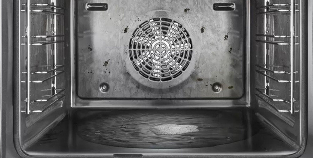 Wnętrze piekarnika z widocznym wentylatorem na tylnej ścianie i metalowymi prowadnicami po bokach. Na dnie piekarnika znajduje się niewielka ilość wody.