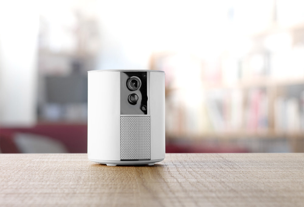 Na drewnianym blacie stoi biały, cylindryczny system alarmowy z kamerą i głośnikiem. W tle widoczne jest rozmyte wnętrze pomieszczenia.