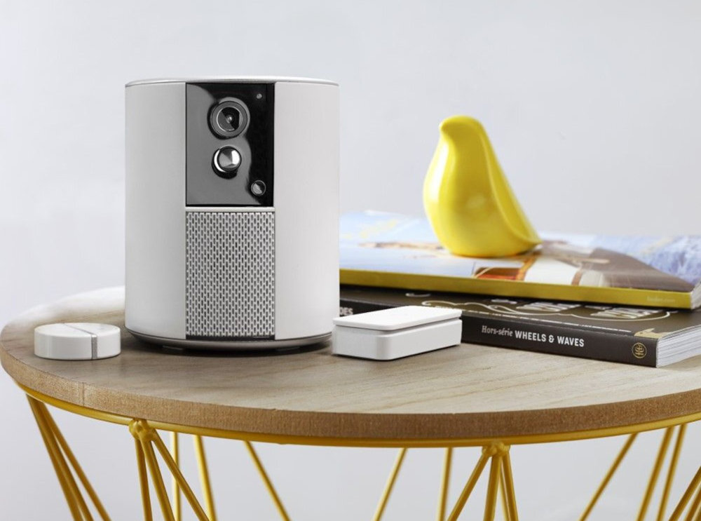 Na okrągłym stoliku znajduje się biały, cylindryczny system alarmowy z kamerą i głośnikiem. Obok leżą dwa białe, prostokątne urządzenia oraz książka z żółtą figurką ptaka na wierzchu.