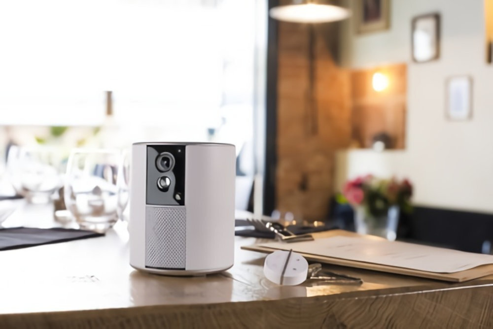 Na drewnianym blacie stoi biały, cylindryczny system alarmowy z kamerą. W tle widoczne są rozmyte elementy wnętrza, takie jak lampy i dekoracje.