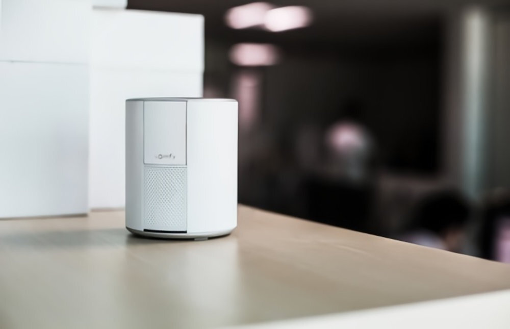 Na drewnianym blacie stoi biały, cylindryczny system alarmowy z widocznym logo \'somfy\'. W tle widać rozmyte wnętrze pomieszczenia.