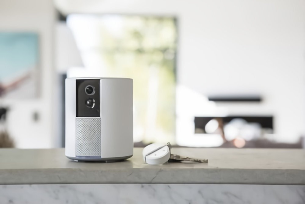 Na blacie stoi biały, cylindryczny system alarmowy z kamerą. Obok leżą klucze i małe urządzenie elektroniczne.