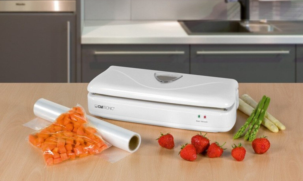 Na blacie kuchennym znajduje się biała zgrzewarka z widocznym logo \'CLATRONIC\'. Obok leżą świeże truskawki, szparagi oraz folia z zapakowanymi marchewkami.