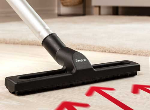 Na podłodze znajduje się czarna ssawka odkurzacza z widocznym logo \'Amica\'. W tle widać fragment dywanu, a na podłodze są czerwone strzałki skierowane w górę.