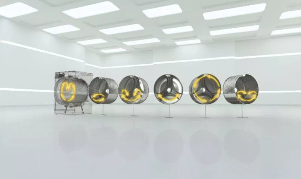 Pięć bębnów pralki ustawionych w rzędzie w jasnym pomieszczeniu, z żółtymi elementami wewnątrz każdego z nich. Pomieszczenie ma nowoczesny wygląd z prostokątnymi panelami na suficie.