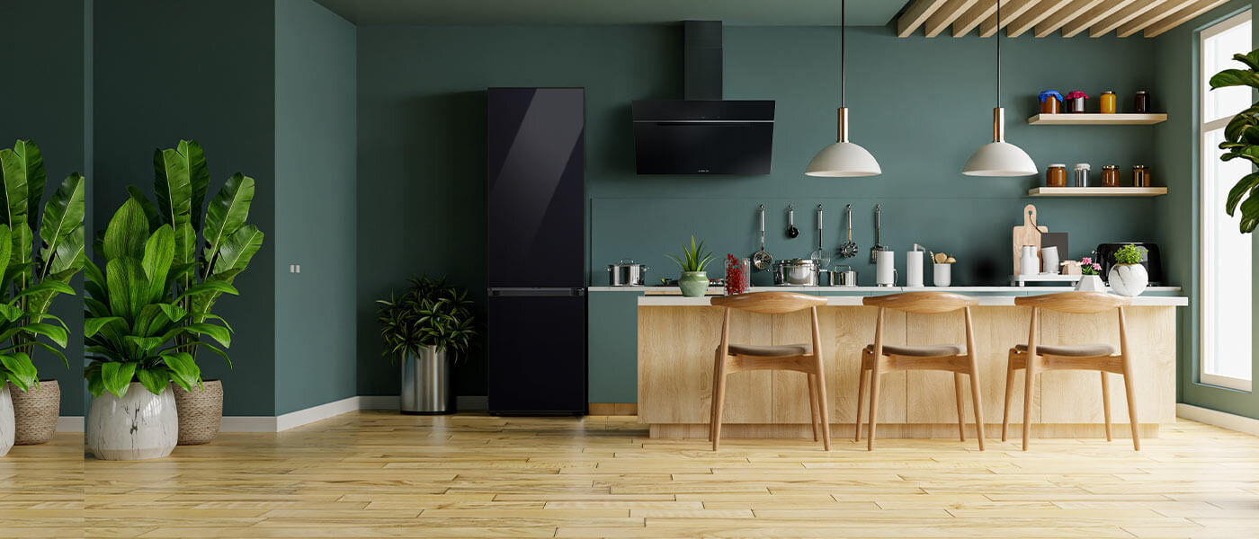 Wnętrze kuchni z czarną lodówką, drewnianym blatem i trzema krzesłami barowymi. Na blacie znajdują się różne akcesoria kuchenne, a w tle widać zielone rośliny i półki z kolorowymi pojemnikami.