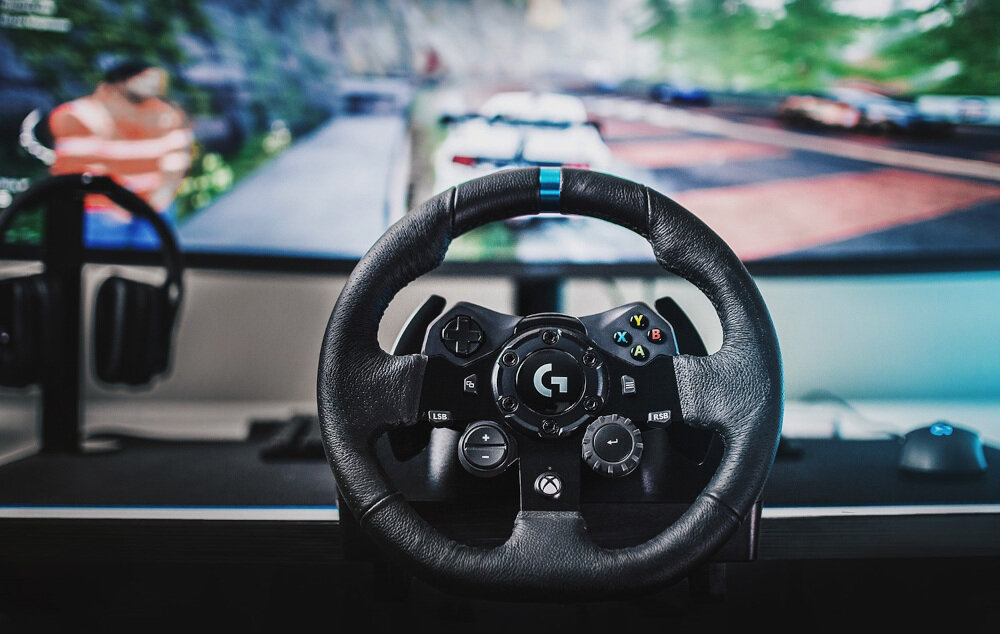 Na pierwszym planie znajduje się kierownica z logo \'G\' na środku, z przyciskami i pokrętłami. W tle widoczny jest ekran z grą wyścigową.