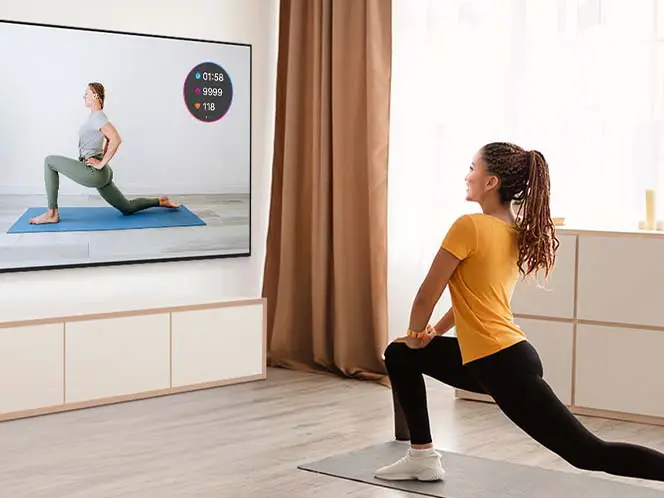 Kobieta ćwiczy, oglądając trenera fitness na telewizorze. Jej statystyki treningu z Galaxy Watch pojawiają się jako nakładka na ekranie.