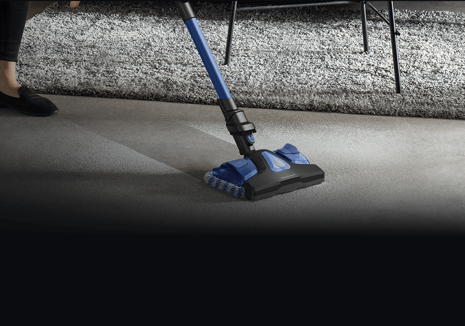 Na obrazie widoczny jest odkurzacz z niebieską rurą i czarną głowicą czyszczącą, używany na dywanie i podłodze. W tle znajduje się fragment dywanu i nogi krzesła.