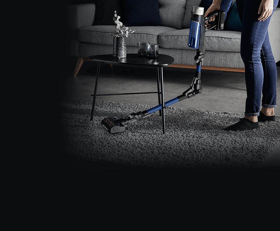 Osoba używa odkurzacza z elastyczną rurą na dywanie w salonie, obok stolika z wazonem i szklanką. W tle widoczna jest szara sofa z poduszkami.