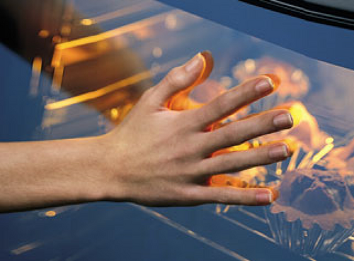 Dłoń dotykająca szklanych drzwi piekarnika, za którymi widać rozgrzane wnętrze z piekącymi się potrawami.