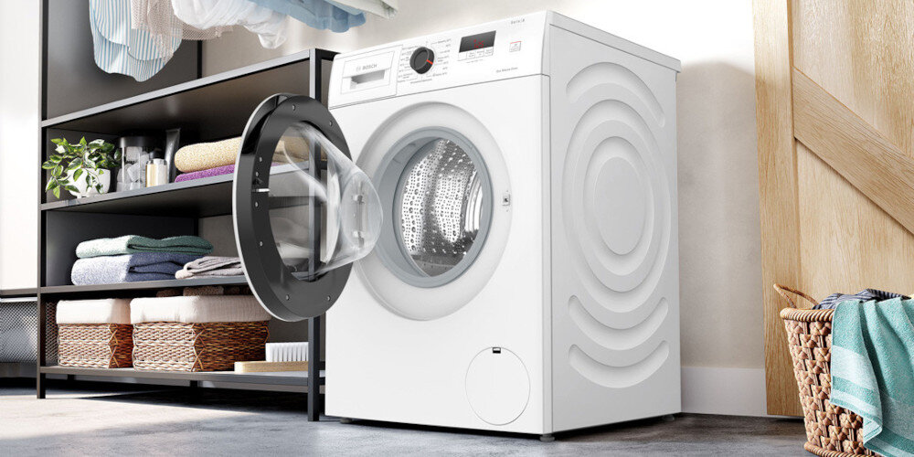 Na obrazie znajduje się biała pralka z otwartymi drzwiami, ustawiona w jasnym pomieszczeniu. W tle widoczny jest regał z ręcznikami i kosz na pranie.