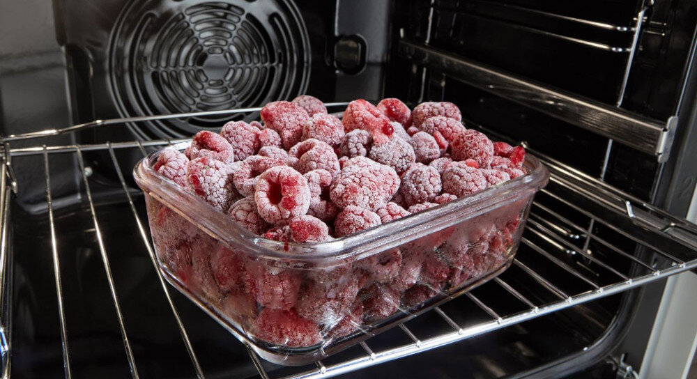 Szklane naczynie wypełnione mrożonymi malinami znajduje się na metalowej półce wewnątrz piekarnika. W tle widoczny jest wentylator piekarnika.