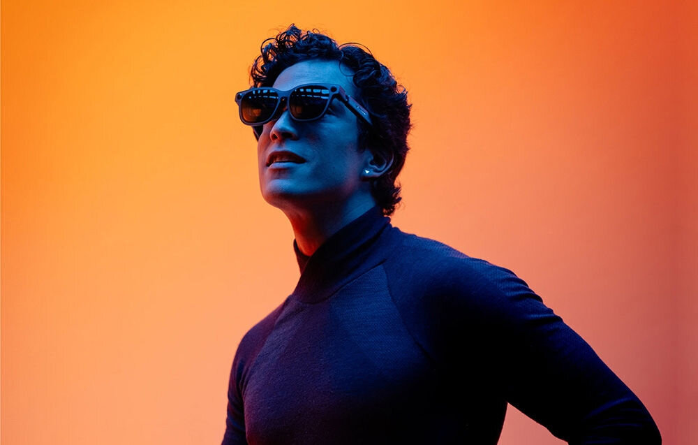 Osoba nosi czarne okulary przeciwsłoneczne na tle gradientu w odcieniach pomarańczowego i niebieskiego. Osoba ma na sobie ciemny golf.