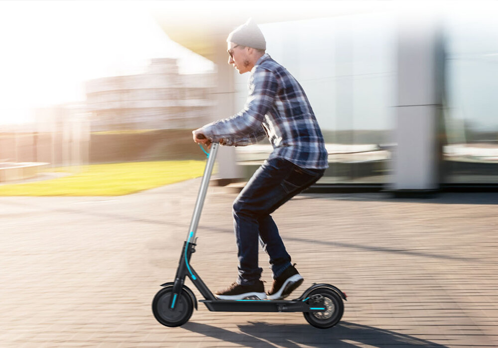 Osoba jedzie na czarno-turkusowej hulajnodze elektrycznej po chodniku. W tle widać rozmyty budynek i trawnik.