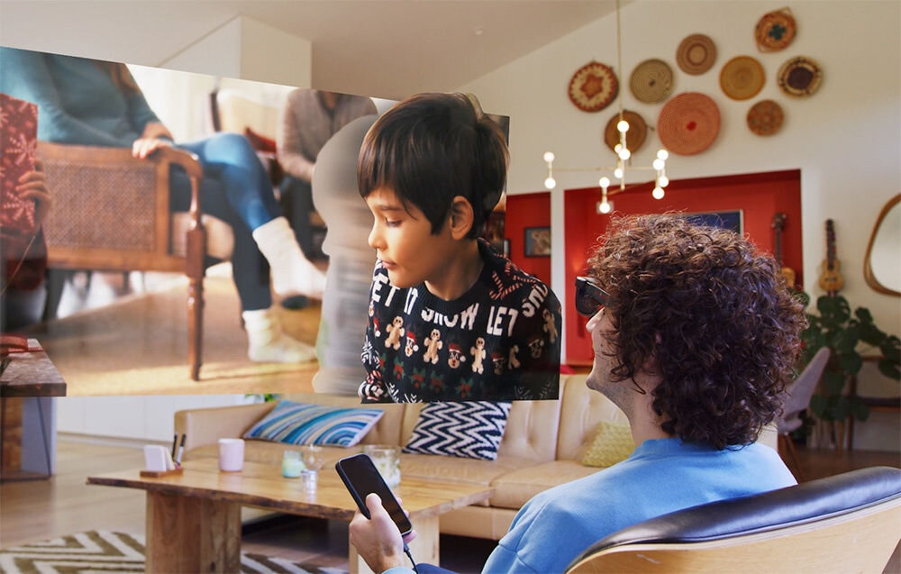 Osoba siedzi w salonie, trzymając w ręku urządzenie, a przed nią widoczny jest obraz dziecka na ekranie. W tle znajduje się nowoczesne wnętrze z dekoracjami na ścianie i meblami.