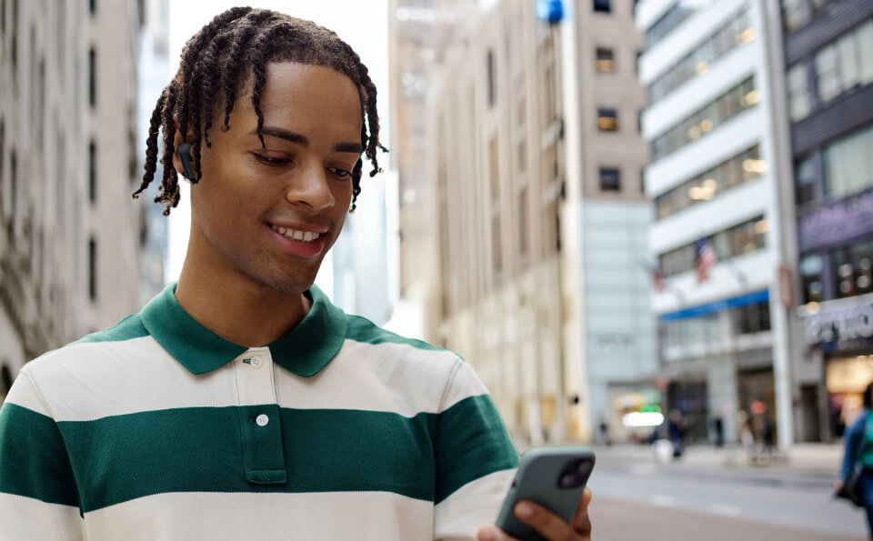 Osoba stoi na ulicy w mieście, trzymając w ręku smartfon. W uszach ma czarne słuchawki dokanałowe.