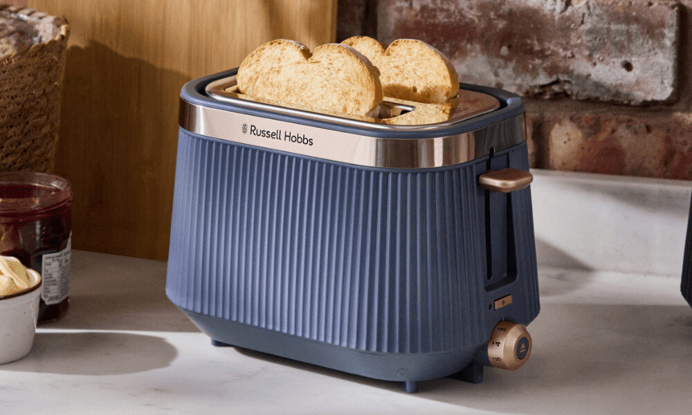Niebieski toster z logo \'Russell Hobbs\' na blacie kuchennym, z dwoma kromkami chleba w środku. W tle widoczne są ceglana ściana i drewniana deska.