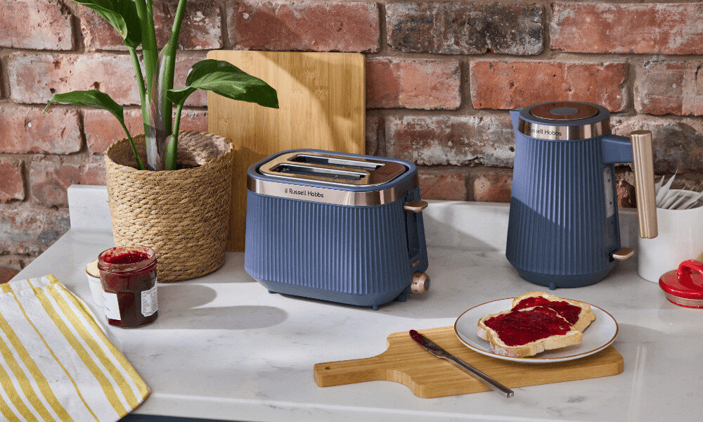 Na blacie kuchennym stoi niebieski czajnik i toster marki Russell Hobbs. Obok znajduje się słoik z dżemem i talerz z kromką chleba posmarowaną dżemem.