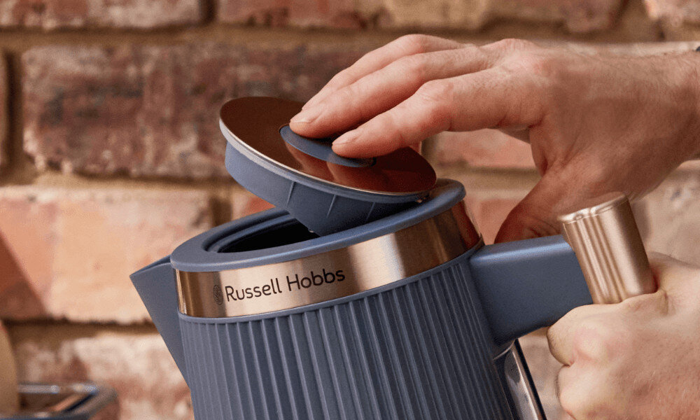 Osoba otwiera pokrywkę niebieskiego czajnika z napisem \'Russell Hobbs\' na tle ceglanej ściany.