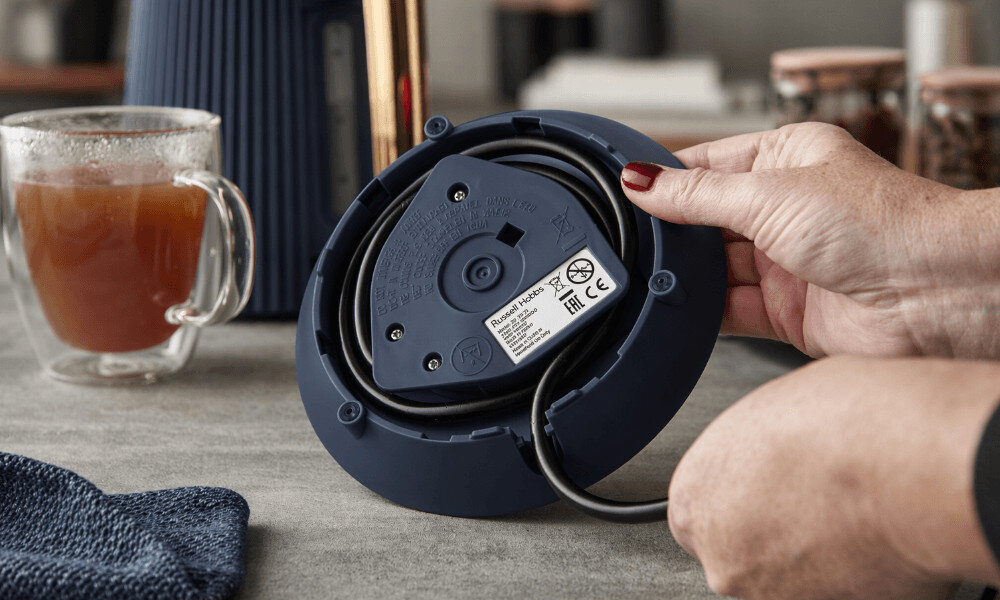 Osoba trzyma podstawę czajnika z nawiniętym kablem, na której widoczne jest logo \'Russell Hobbs\'. Obok stoi szklanka z napojem.